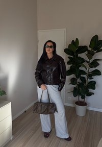 Žena s dlhými tmavými vlasmi, oblečená v čiernej koženej bunde, šedých širokých nohaviciach, so slnečnými okuliarmi, ktorá drží hnedú kabelku, stojí v miestnosti s rastlinami.