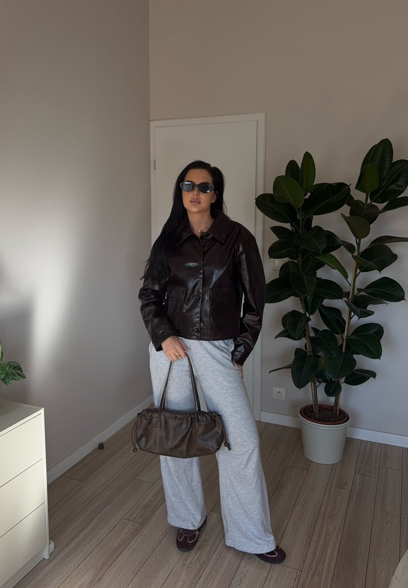 Frau mit langen dunklen Haaren, die eine schwarze Lederjacke, graue weite Hosen und Sonnenbrillen trägt, mit einer braunen Handtasche in der Hand, steht in einem Raum mit Pflanzen.