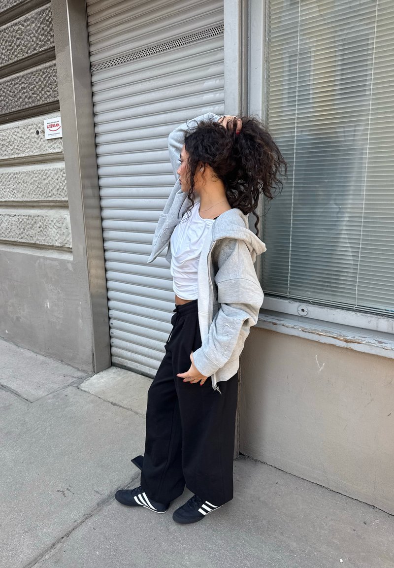 Femme en vêtements décontractés se tient sur le trottoir, ajustant ses cheveux bouclés, à côté d'un volet métallique fermé et d'une fenêtre avec des stores.