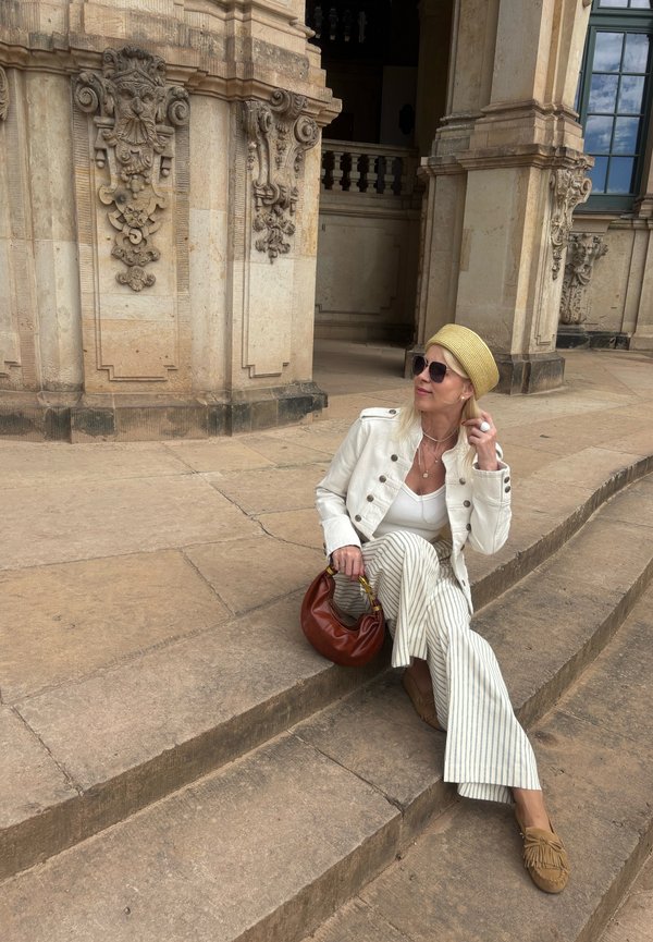 Femme en veste blanche, pantalon rayé et béret beige assise sur des marches en pierre près d'une façade classique ornée de détails sculptés.