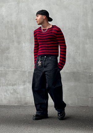 Suéter de punto a rayas rojo y azul marino, jeans negros holgados con bolsillos laterales, combinados con zapatos negros gruesos y collares de plata en capas.