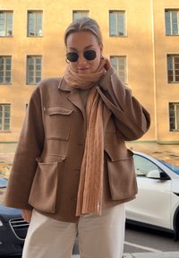 Brauner Wollmix Oversized-Jacke mit zwei vorderen Taschen, kombiniert mit einem strukturierten hellbeigen Schal. Eine Sonnenbrille rundet das Outfit ab.