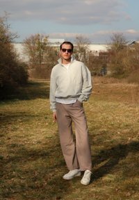 Svetlo šedá mikina na zips, svetlo modrá košeľa pod ňou, béžové široké nohavice a biele tenisky. Hnedé vlasy a slnečné okuliare. Vonkajšie prostredie.
