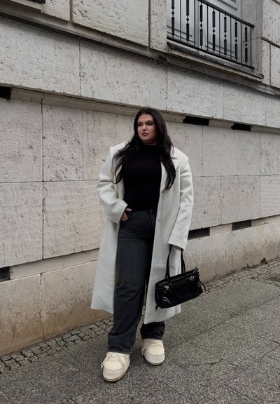 Abrigo largo de textura blanca, top negro ajustado, vaqueros oscuros, zapatos de color claro y un pequeño bolso negro. Entorno urbano con pared de piedra.