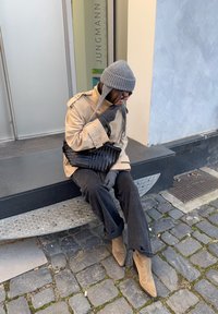 Beżowy trencz, szary dziergany kapelusz, ciemne rękawiczki i rozszerzane czarne spodnie. Czarna pleciona torba i beżowe botki na obcasie. Siedzi na stopniu.