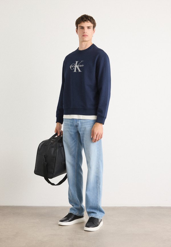 Sweatshirt azul-marinho com o logótipo da Calvin Klein, jeans azul-claro, mala de couro preta e ténis pretos com soles brancos e detalhes em costura.