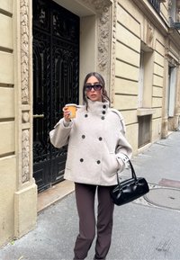 Jasnoszary dwurzędowy płaszcz z dużymi czarnymi guzikami, noszony na brązowych spodniach, trzymany z czarną torebką i kubkiem kawy.