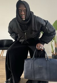 Čierna bomber bunda, čierny šál, nadmerné čierne nohavice, drží tmavomodrú koženú tašku so striebornými doplnkami. Na pozadí drevená stolička a rastlina.