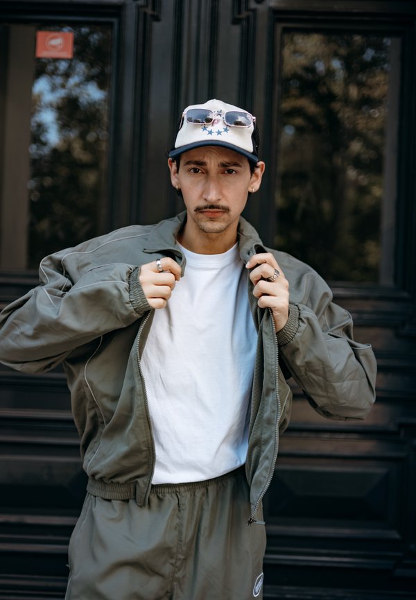 Olivgrön bomberjacka med dragkedja fram, vit t-shirt under, matchande byxor och en keps med solglasögon på skärmen.