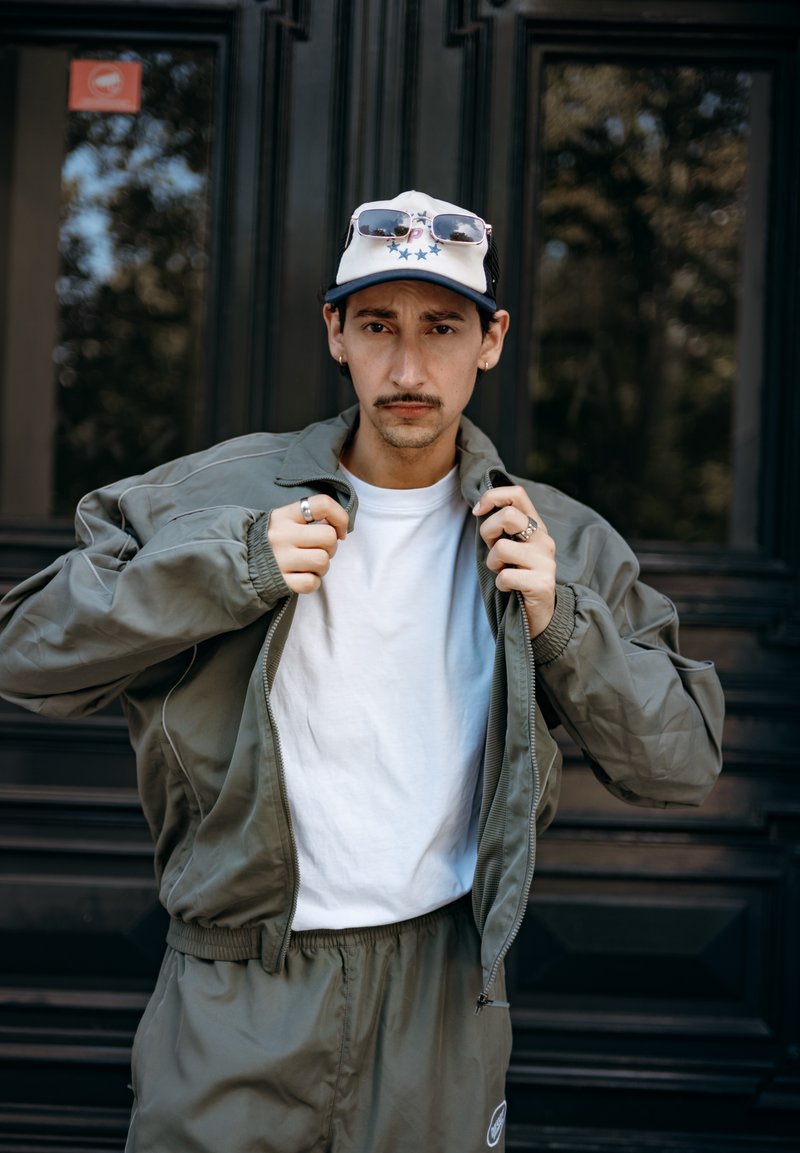 Olivgrüne Bomberjacke mit Reißverschluss vorne, weißes T-Shirt darunter, passende Hose und eine Kappe mit Sonnenbrille am Schirm.