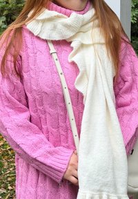 Άτομο που φοράει ροζ πουλόβερ με πλεκτό ανάγλυφο σχέδιο και λευκό κασκόλ με βολάν, κρατώντας έναν λευκό ι μάντα έξω, με φόντο πράσινο τοπίο.