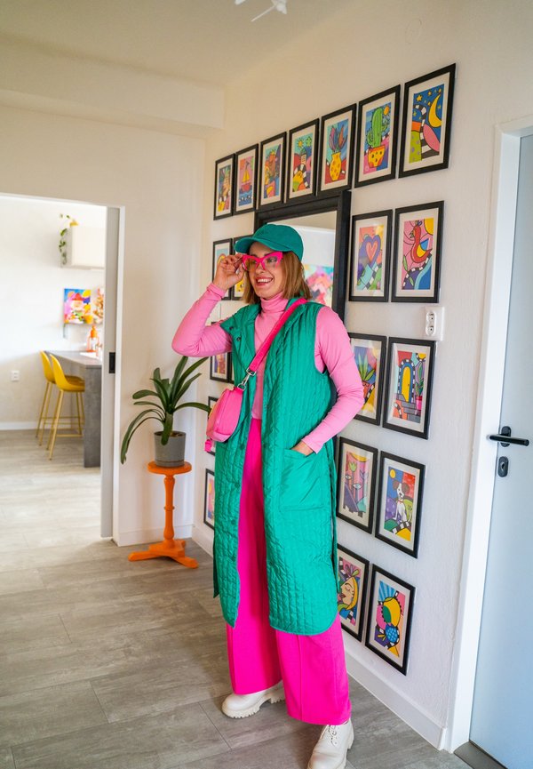 Femme en tenue vive rose et verte ajustant des lunettes roses, debout près d’un mur avec des œuvres d’art encadrées colorées dans un intérieur domestique lumineux et moderne.