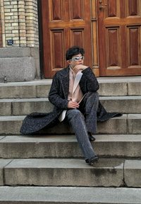 Palton texturat negru cu guler lung, cămașă bej, pantaloni cu un model strălucitor și ochelari de soare reflectanți, așezat pe trepte de piatră. Uși din lemn în fundal.