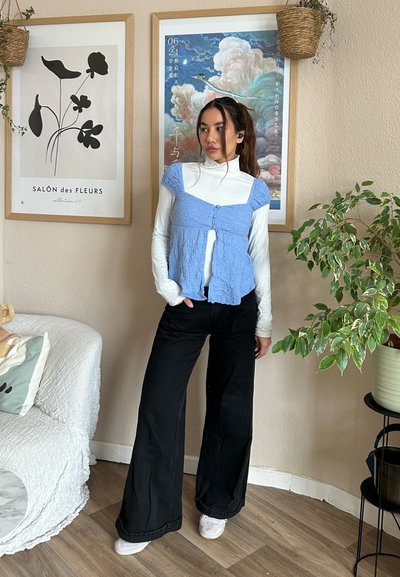 Top de tela arrugada en azul claro, superpuesto a un cuello de tortuga blanco; combinado con pantalones negros de pierna ancha y zapatillas blancas. Entorno interior con plantas.