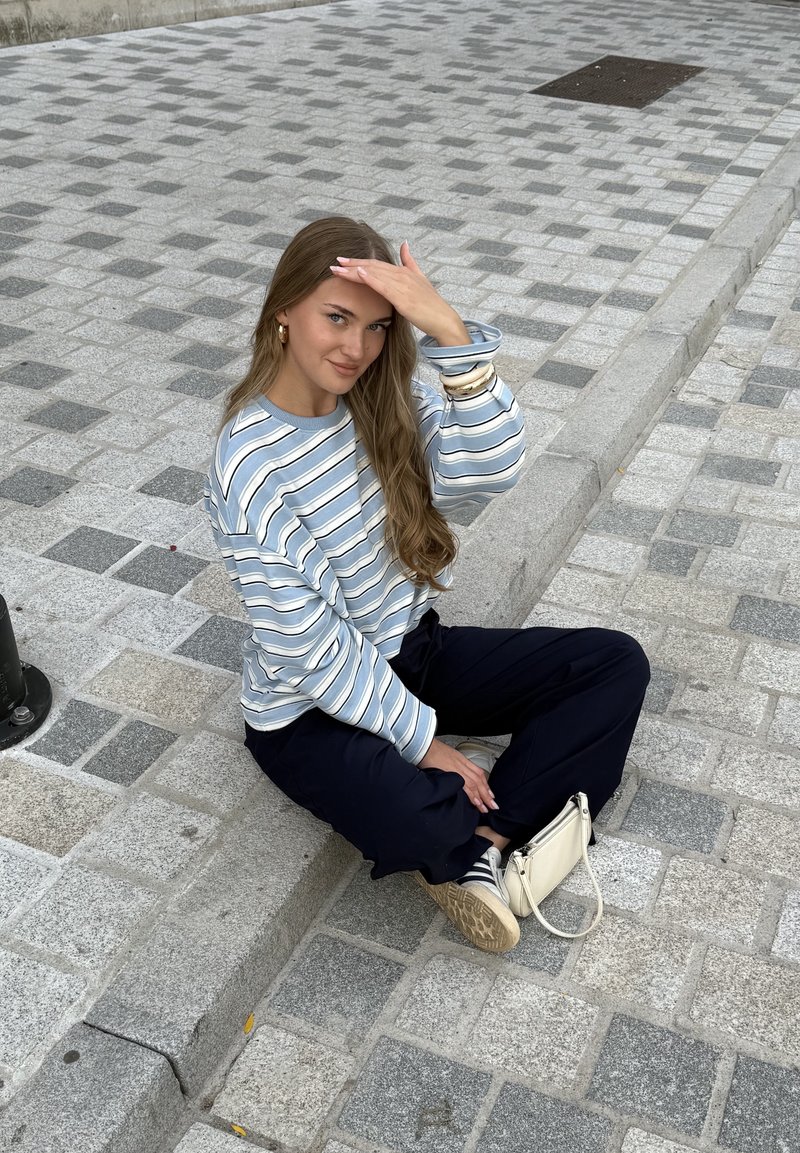 Camisa de manga longa azul e branca com riscas, calças pretas e ténis claros. Os acessórios incluem pulseiras e uma pequena bolsa branca.