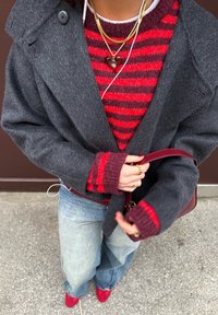 Manteau en laine gris à larges revers, porté sur un pull rayé rouge et bordeaux. Jean et chaussures rouges. Accessoires comprenant un collier avec pendentif en forme de cœur.