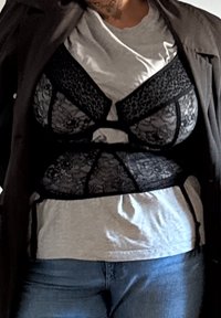 Bralette en dentelle noire sur un t-shirt gris, présentant un motif floral et un design structuré, superposé sous une veste sombre.