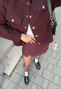 Jachetă din lână bordeaux cu nasturi aurii, bluză albă, fustă bordeaux, mocasini negri și șosete albe; stând pe o suprafață pavată gri.