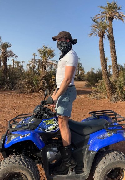 Vehículo todoterreno azul con neumáticos robustos, detalles en negro y un diseño gráfico. Persona con una camiseta blanca y pantalones cortos de mezclilla se encuentra al lado.