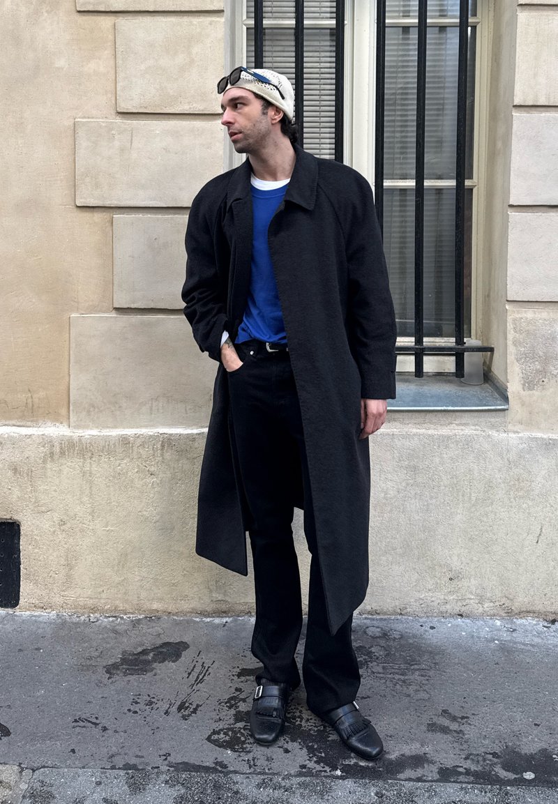 Homme portant un long manteau noir, une chemise bleue, un pantalon noir, des chaussures noires et un bonnet blanc, debout dans la rue à côté d'un mur beige et d'une fenêtre avec des barreaux, regardant vers la gauche.