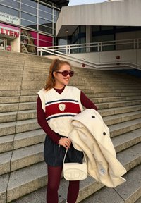 Vestă albă tricotată cu dungi roșii și albastre peste un top cu mâneci lungi vișiniu, asortată cu pantaloni scurți cu dungi, ținând o geacă crem și o geantă.