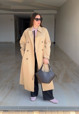 Mujer con gabardina beige, pantalones negros de pierna ancha, zapatos rosas, gafas de sol, sosteniendo un bolso negro en un pasillo.