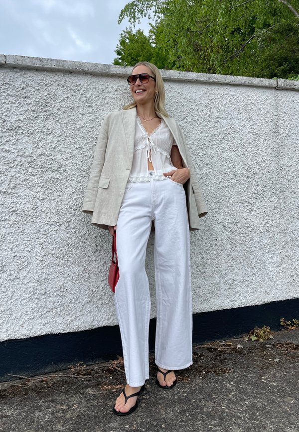 Lachende vrouw met een zonnebril, beige blazer, witte cropped blouse, witte wijde broek, zwarte slippers en een rode handtas vasthoudend buiten.