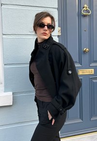 Veste noire surdimensionnée en tissu texturé, avec un col haut, de gros boutons et une coupe décontractée, portée avec un haut marron et un pantalon noir.