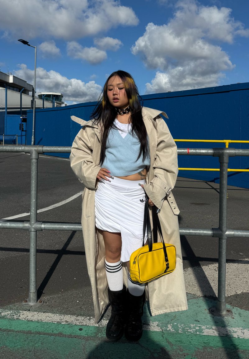 Une femme pose près d’une balustrade, vêtue d’un trench beige, d’un haut bleu, d’une jupe blanche, de chaussettes rayées et de bottes noires. Elle tient un sac jaune vif.