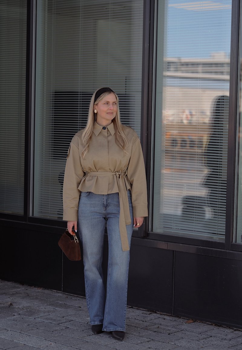 Femme aux cheveux blonds longs portant une veste ceinturée beige, un jean bleu, un serre-tête noir, tenant un sac à main marron, debout près de fenêtres en verre en plein air.