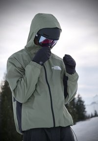 Personne portant une veste verte The North Face, des gants noirs et un casque de ski ajustant ses lunettes dans un environnement enneigé en plein air.