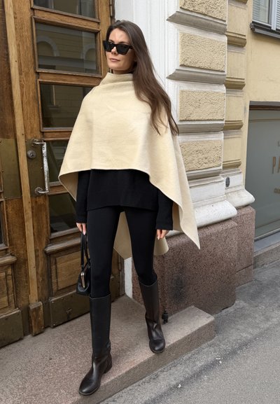 Mujer con capa beige, suéter negro, leggings, botas hasta la rodilla y gafas de sol, parada fuera de un edificio junto a una puerta de madera.