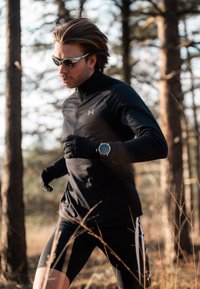 Haut de course noir avec col montant, gants noirs et shorts noirs à rayures blanches. Porte des lunettes de soleil et une montre intelligente.