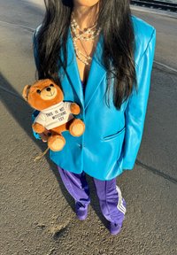 Saco de imitación cuero azul brillante sobre pantalones de chándal morados, sosteniendo un oso de peluche marrón que lleva una camiseta blanca con texto negro.