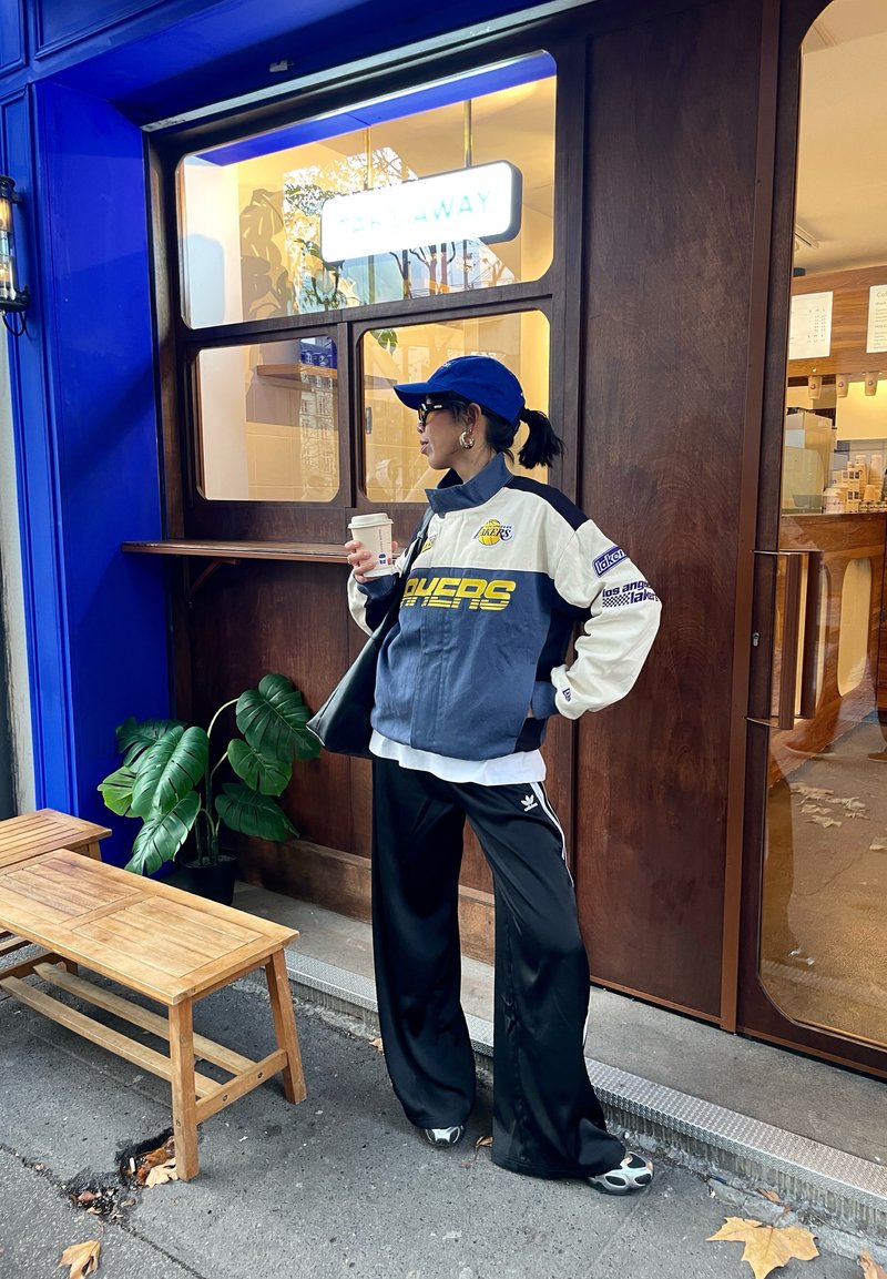 Veste de survêtement bleue et blanche avec logo, pantalon de survêtement noir et casquette noire. Tenant une tasse de café, debout près d'un banc en bois et de plantes.