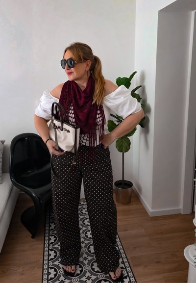 Mujer con gafas de sol, top blanco con hombros descubiertos, bufanda granate y pantalones negros de lunares, sosteniendo un bolso blanco en una sala de estar moderna.