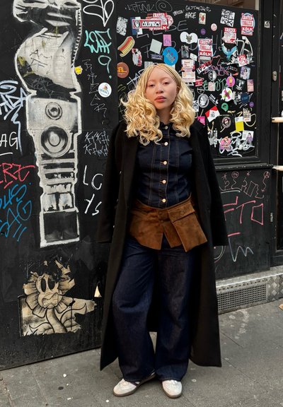 Camisa de mezclilla con costuras claras, cinturón de ante marrón, pantalones vaqueros de pierna ancha, abrigo largo negro y zapatillas blancas frente a una pared cubierta de graffiti.
