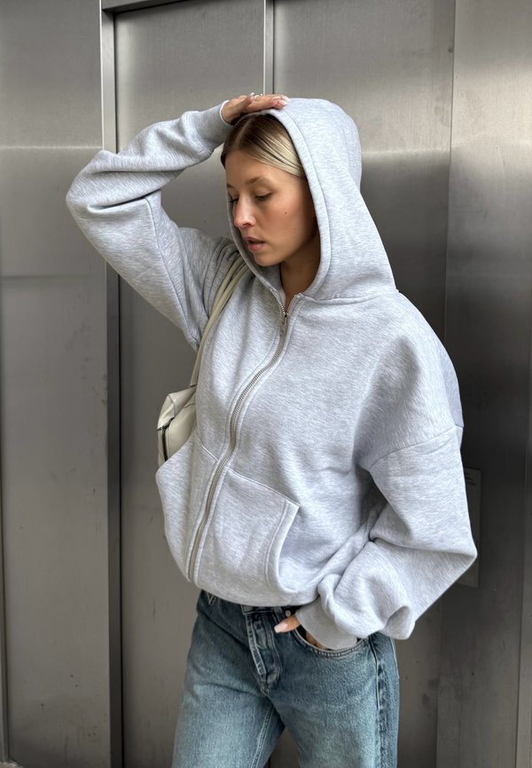 Grå zip-up hættetrøje i fleece materiale med to frontlommer, båret med højtaljede lyseblå jeans og en skuldertaske.