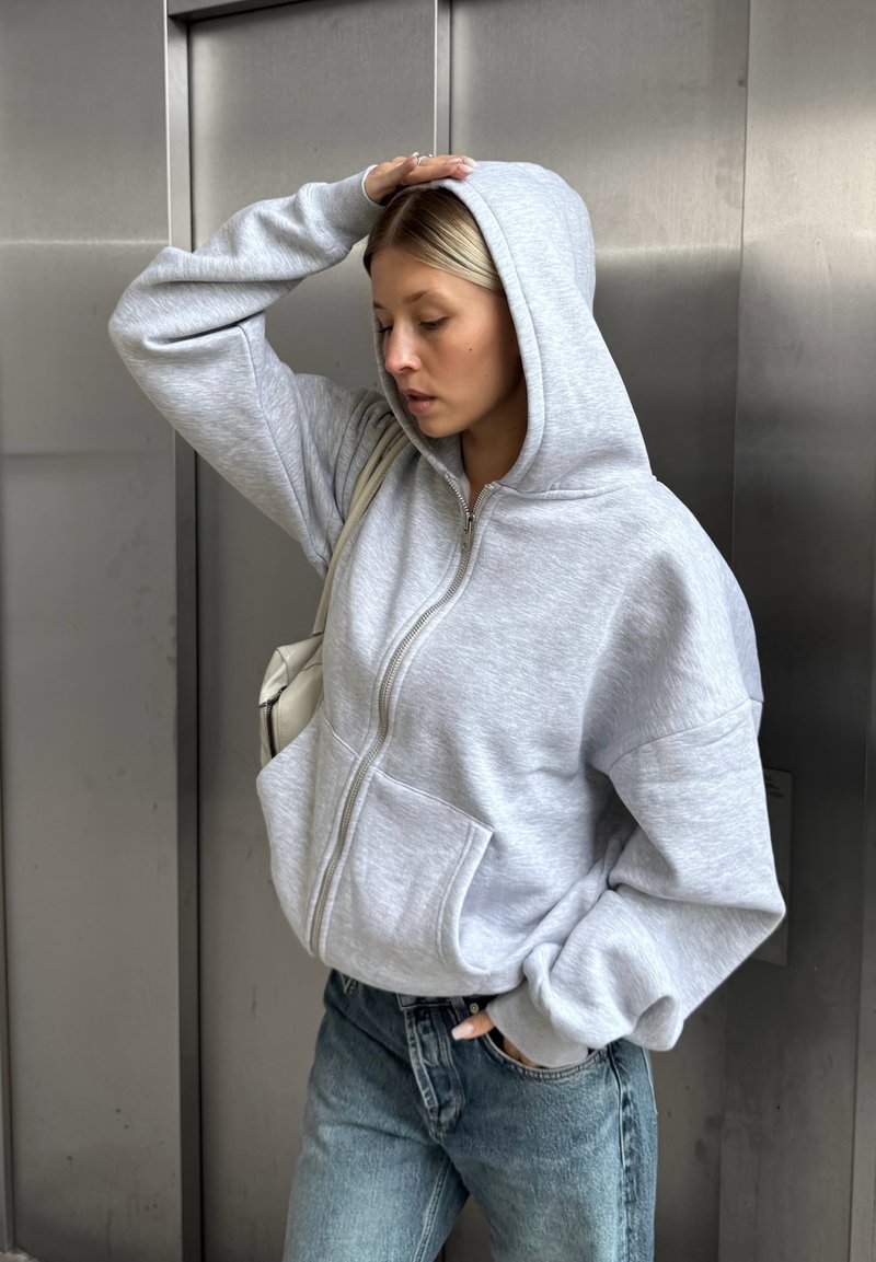 Grauer Reißverschluss-Hoodie aus Fleece mit zwei Fronttaschen, getragen mit hoch taillierten hellblauen Jeans und einer Umhängetasche.