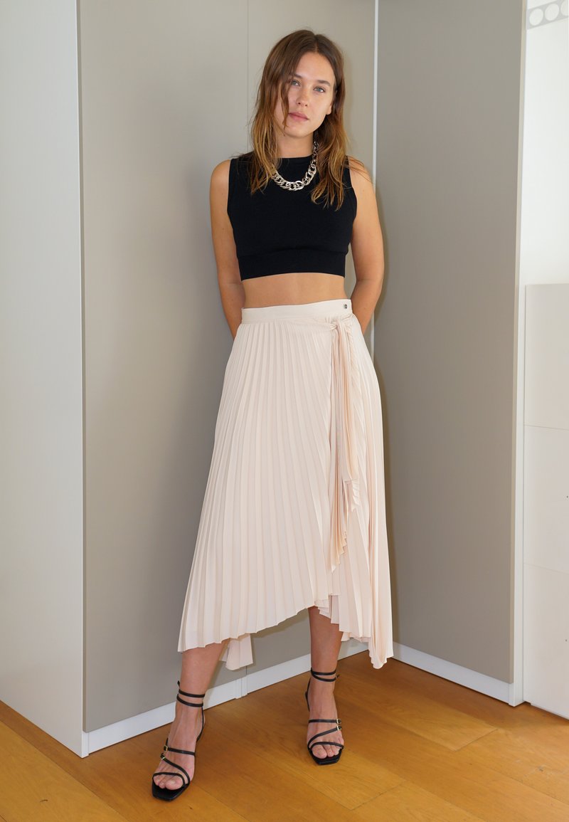 Pleatattu midihame pehmeässä beessissä, jossa on kietaisumalli ja sivusolmu. Musta toppi, hopeaketju kaulakoru ja mustat korkeat korot.