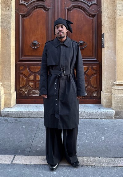 Hombre de pie frente a una puerta de madera, vistiendo un abrigo largo negro con cinturón, pantalones anchos negros, zapatos negros y un turbante negro con un lazo.