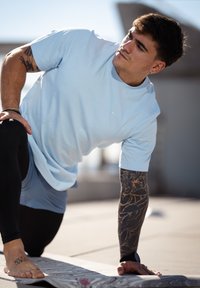 Jasnoniebieska sportowa koszulka z krótkim rękawem i okrągłym dekoltem, zestawiona z czarnymi dopasowanymi legginsami. Charakterystyczne tatuaże na prawej ręce.