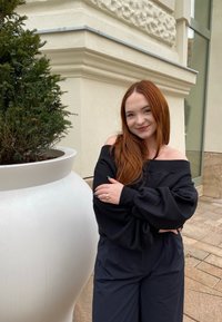 Suéter preto off-shoulder com mangas volumosas, combinado com calças escuras de cintura alta. Vaso redondo branco ao fundo, com folhagem verde visível.
