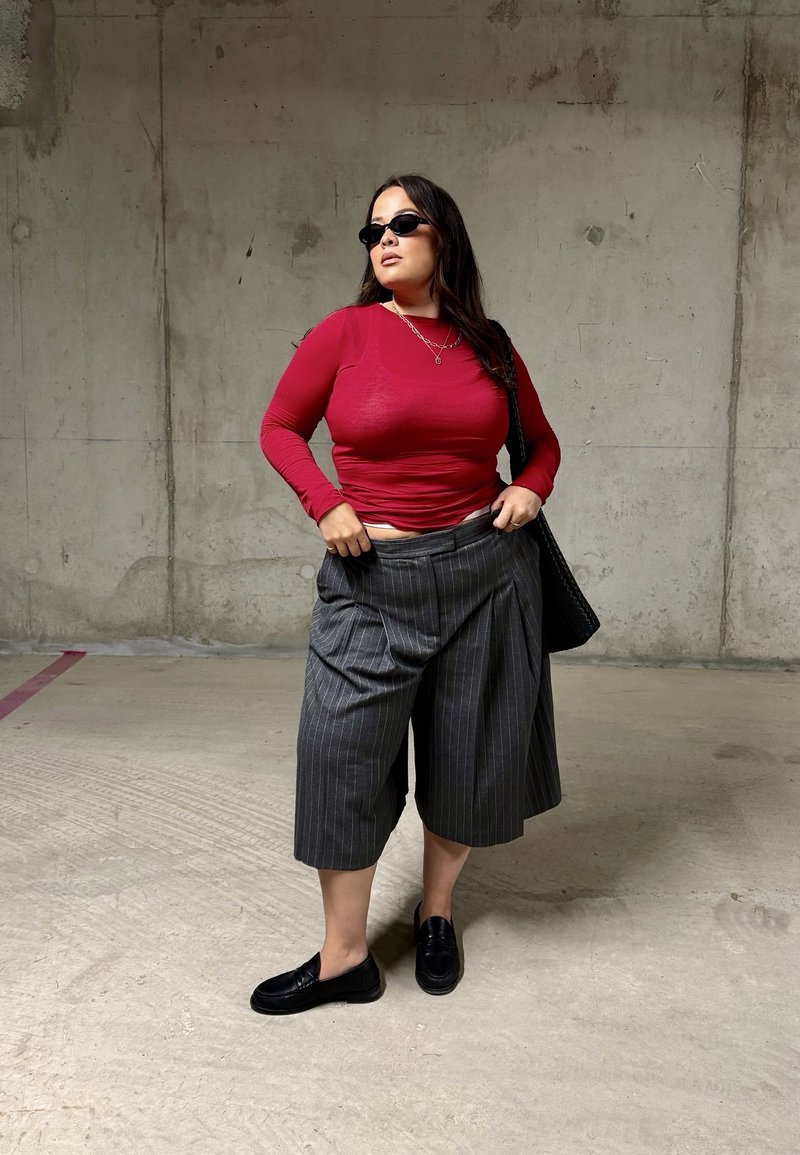 Femme portant des lunettes de soleil noires, un haut rouge à manches longues, un pantalon large gris à fines rayures et des mocassins noirs, se tient contre un mur en béton.