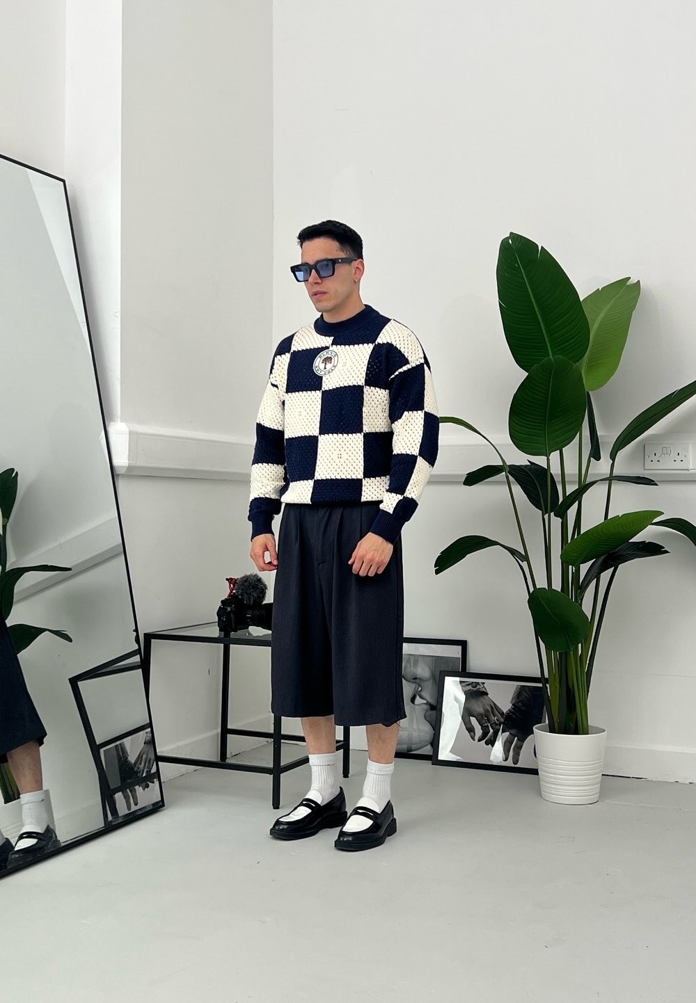 Marineblauw en crèmekleurig geblokt gebreid vest, losse donkere short, witte sokken, zwarte instappers; grote groene plant en spiegel op de achtergrond.
