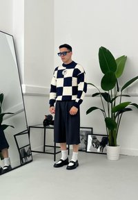 Marineblauw en crèmekleurig geblokt gebreid vest, losse donkere short, witte sokken, zwarte instappers; grote groene plant en spiegel op de achtergrond.