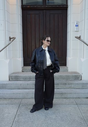 Mujer con chaqueta de cuero negra, camisa blanca, corbata, pantalones anchos negros y gafas de sol, de pie sobre unos escalones de piedra frente a una gran puerta de madera oscura.