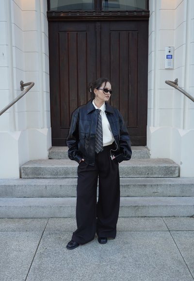 Mujer con chaqueta de cuero negra, camisa blanca, corbata, pantalones anchos negros y gafas de sol, de pie sobre unos escalones de piedra frente a una gran puerta de madera oscura.