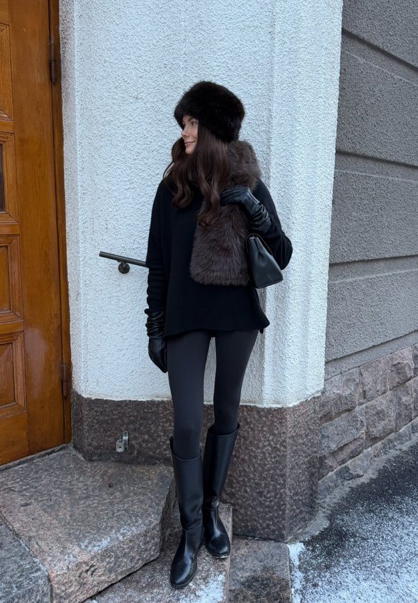 Vrouw die zwarte laarzen, legging, trui, handschoenen en een bontmuts draagt, met een bont sjaal en een zwarte handtas in haar handen, staand bij stenen trappen buiten.