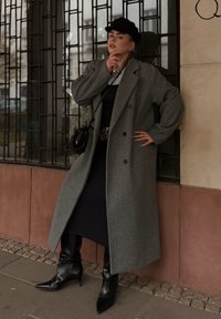 Dlhý sivý vlnený kabát s prehnaným dizajnom, nosený cez čierne šaty. Kombinovaný s čiernymi čižmami na podpätku so špicatou špičkou a čiernou baretou.